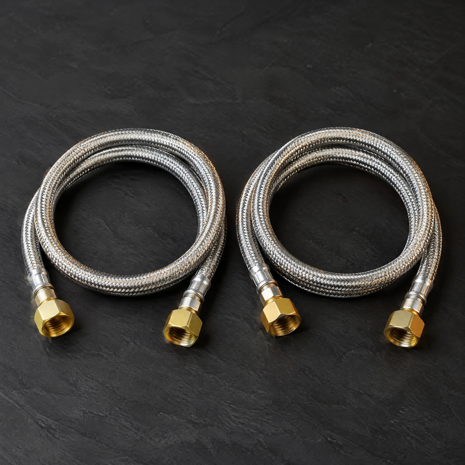 Two coiled braided faucet supply lines with brass-colored nuts on a dark slate surface