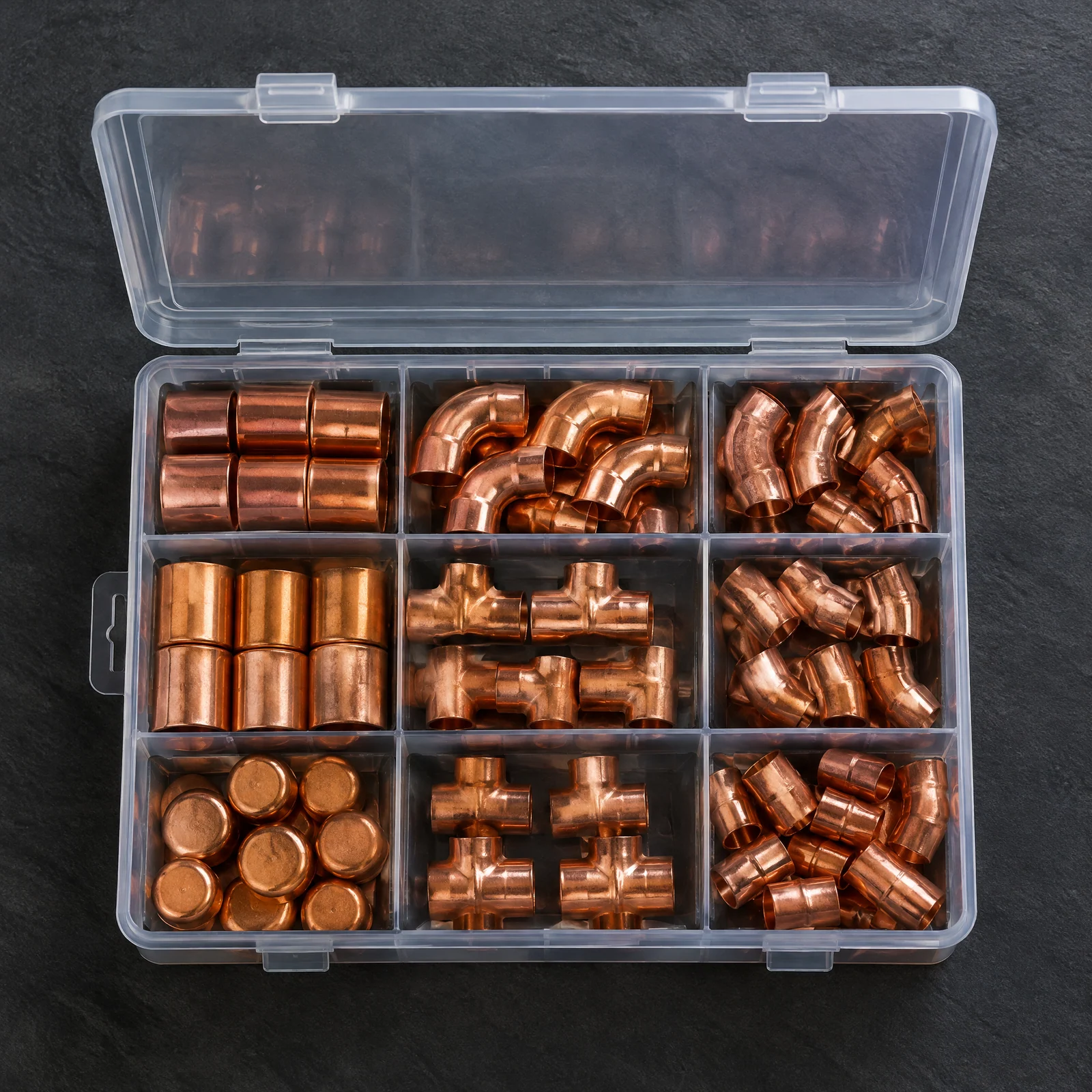 Open clear case with nine compartments of copper-colored couplings, elbows, tees and caps on a dark slate surface