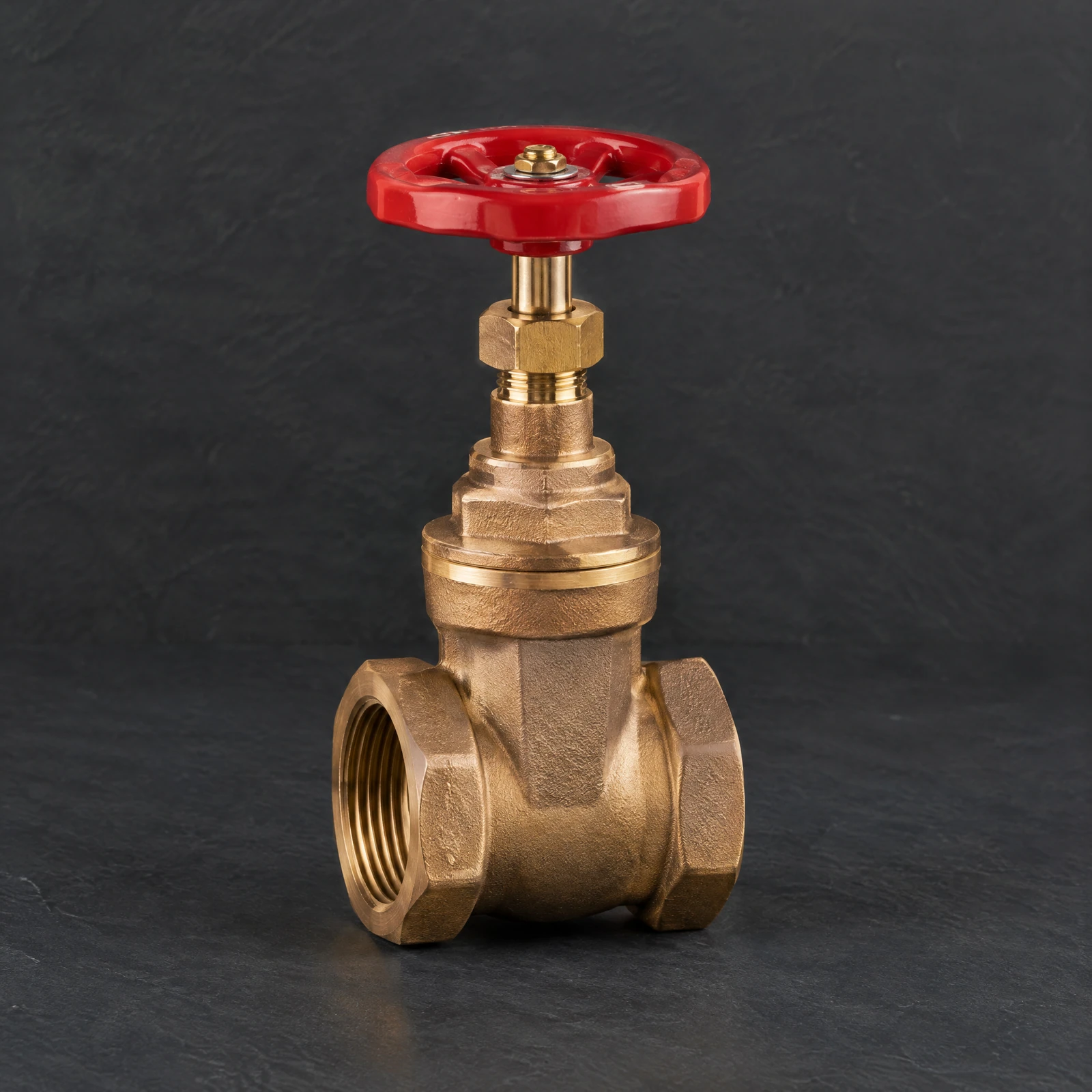 Bronze-toned gate valve with a round red wheel handle on a dark slate surface