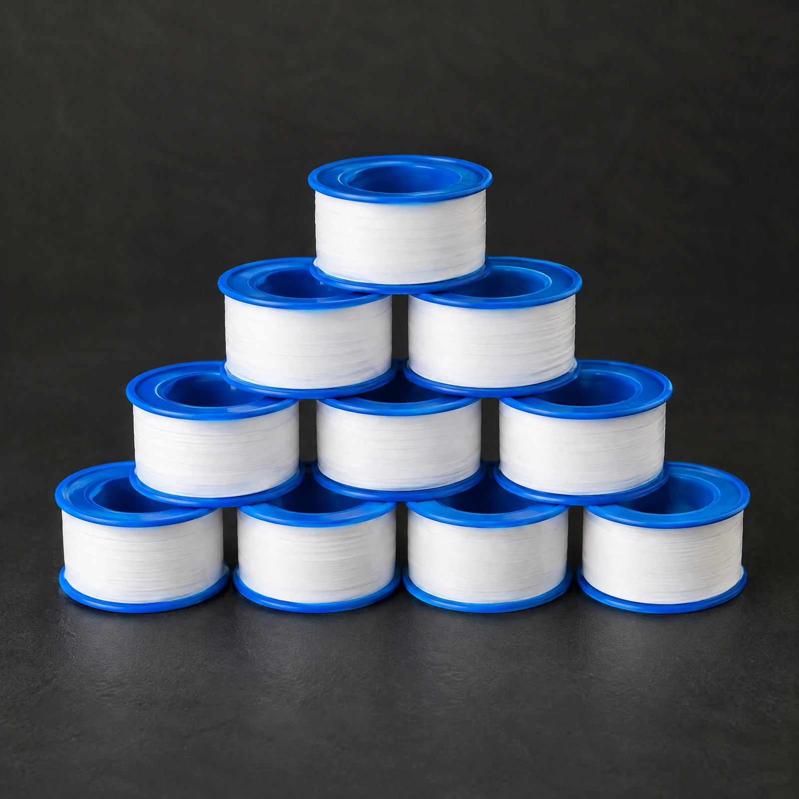 Ten small spools of white thread seal tape on blue cores stacked in a pyramid on a dark slate surface